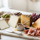 Assorted cheeses, cured meats, and fruits on a marble board with a window in the background.